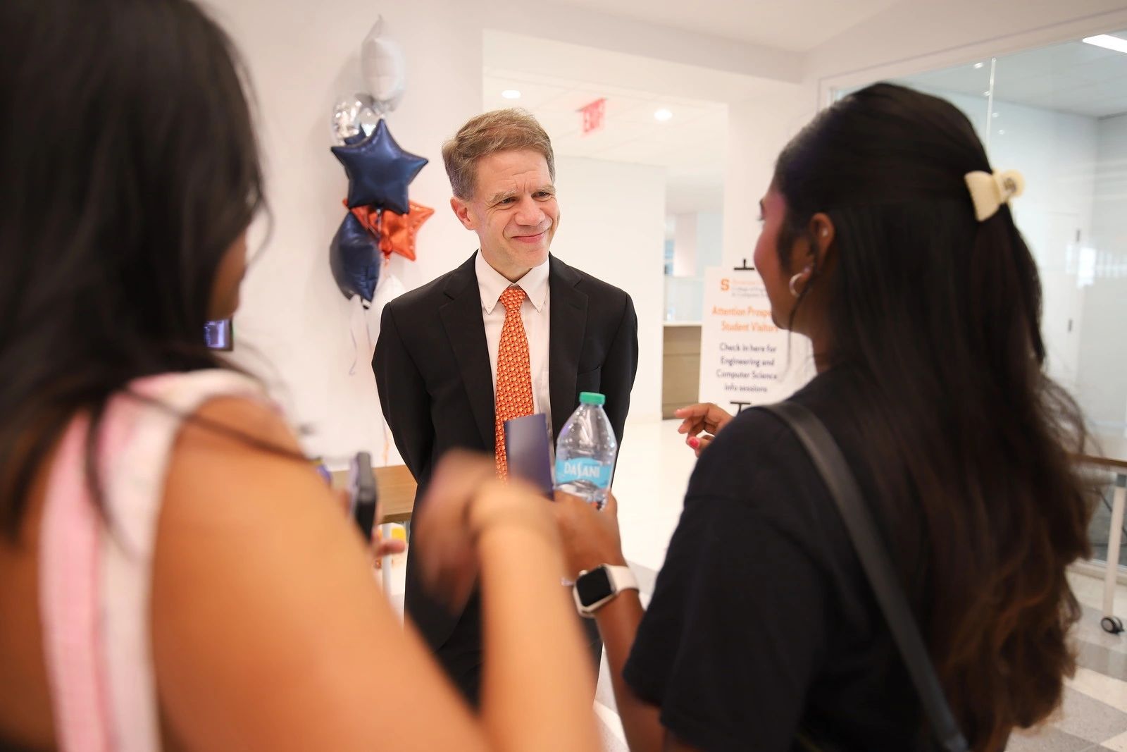 Man in a suit speaking with two individuals indoors, with balloons and a wall sign in the background.