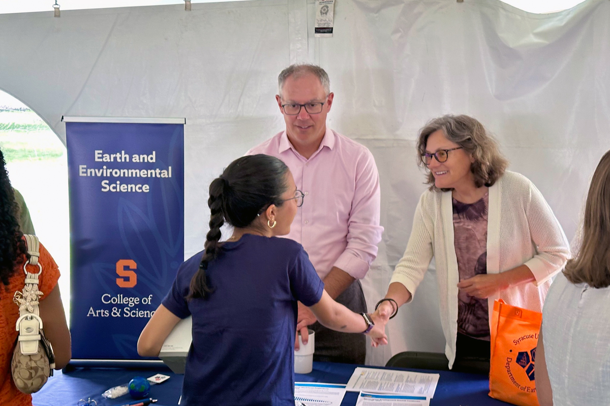 A man in glasses and a pink shirt and a woman in a white cardigan shake hands with a student at an outdoor event. A blue banner reading "Earth and Environmental Science" and "College of Arts & Science" with the Syracuse "S" logo is visible in the background under a white tent.