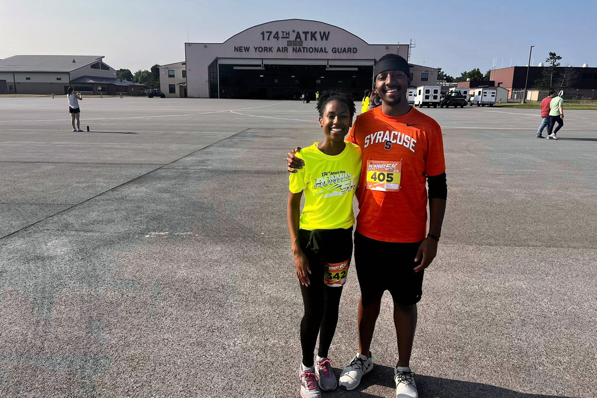 Two smiling runners with race bibs posing together on airport tarmac in front of New York Air National Guard 174th ATKW hangar, wearing bright yellow and orange Syracuse shirts.