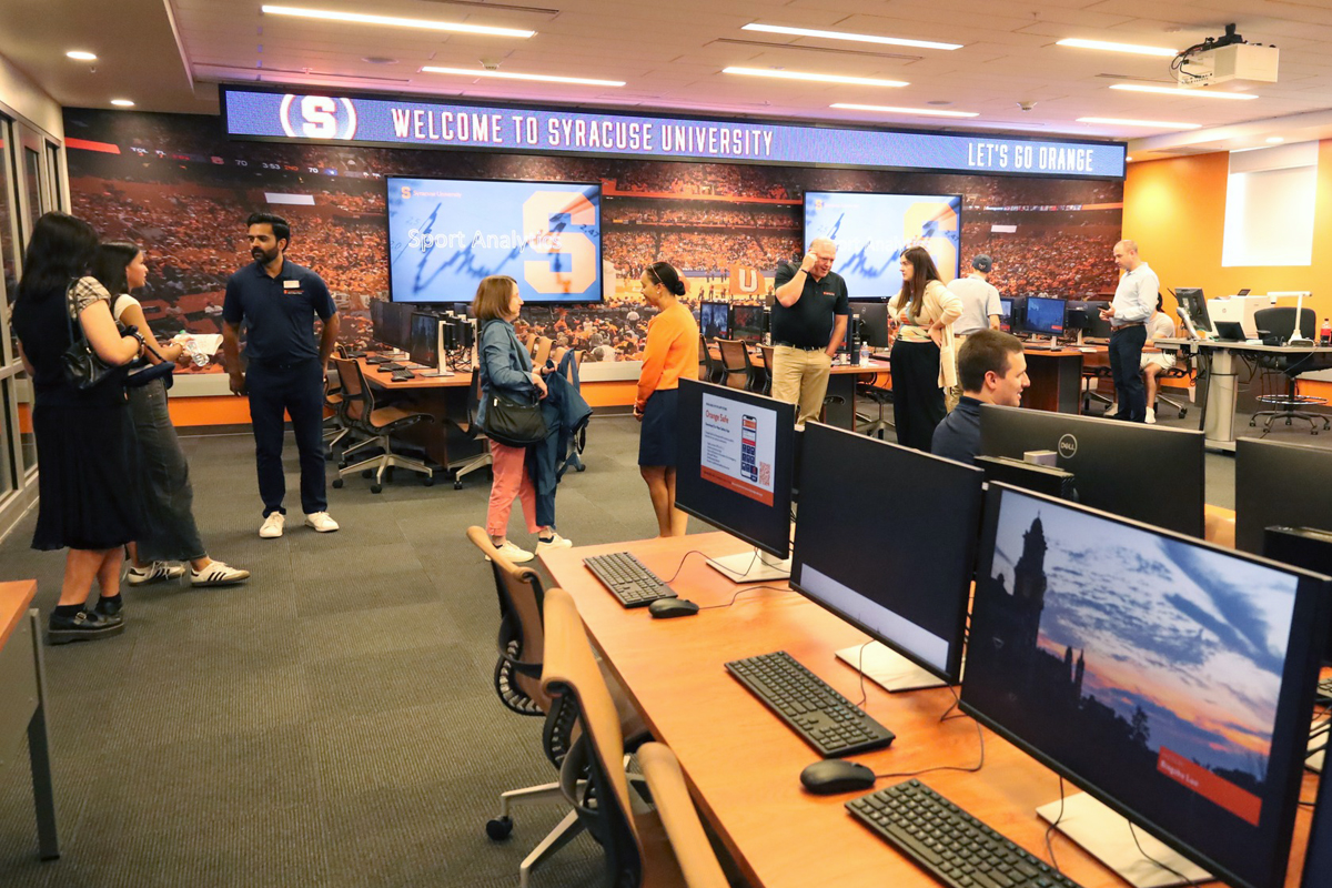 Brightly lit computer lab at Syracuse University with desktop stations, large digital screens displaying welcome messages, and people standing and conversing in a professional, academic setting.