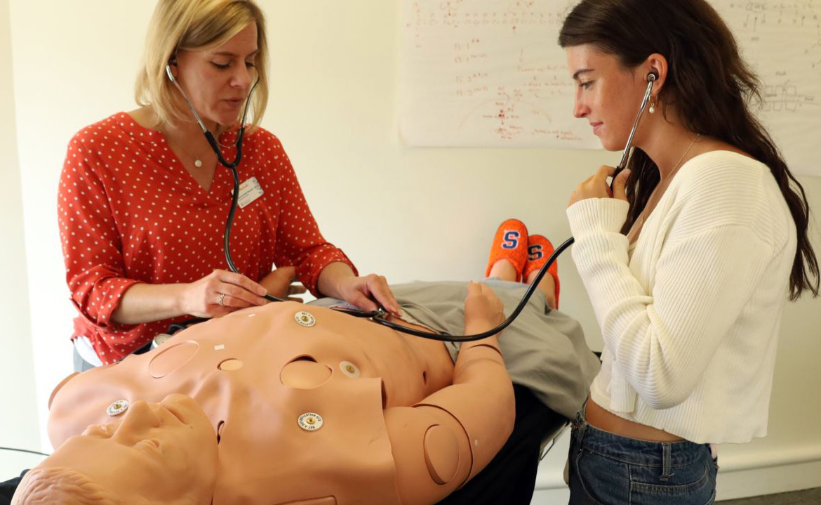 Two people using stethoscopes to examine a medical training mannequin with sensors on its chest and abdomen.
