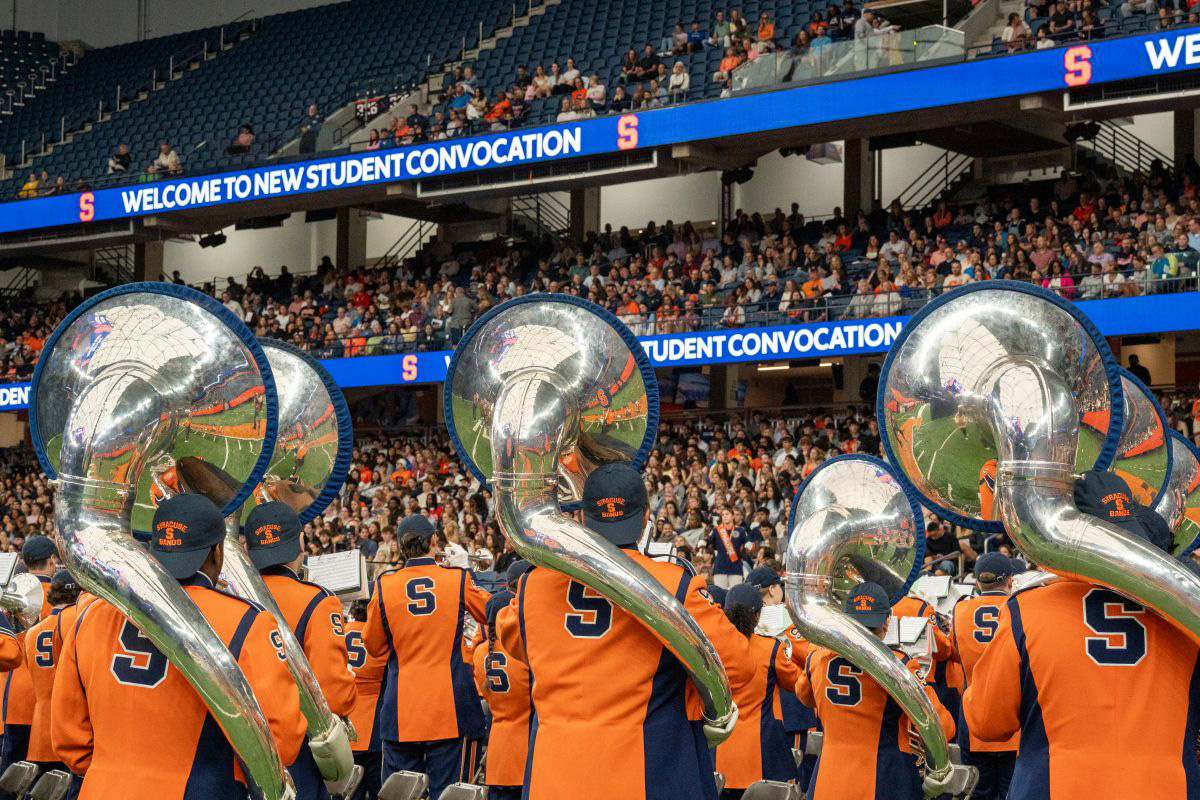 Marching band in orange and blue uniforms performing in a stadium with 'WELCOME TO NEW STUDENT CONVOCATION' displayed above the crowd.