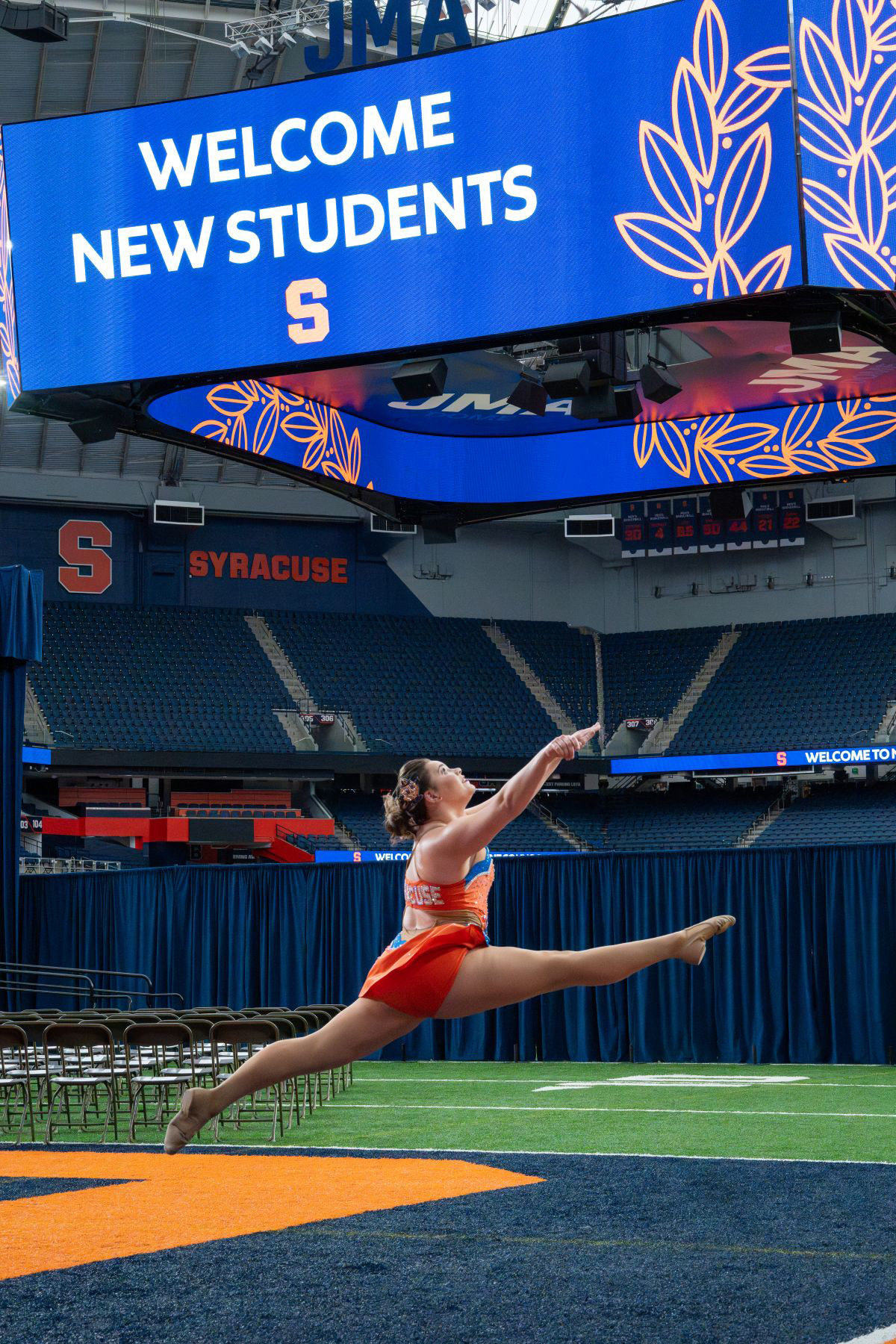 Dancer in orange leaping on the football field in Syracuse’s JMA Wireless Dome, with 'WELCOME NEW STUDENTS' displayed on a large screen at New Student Convocation