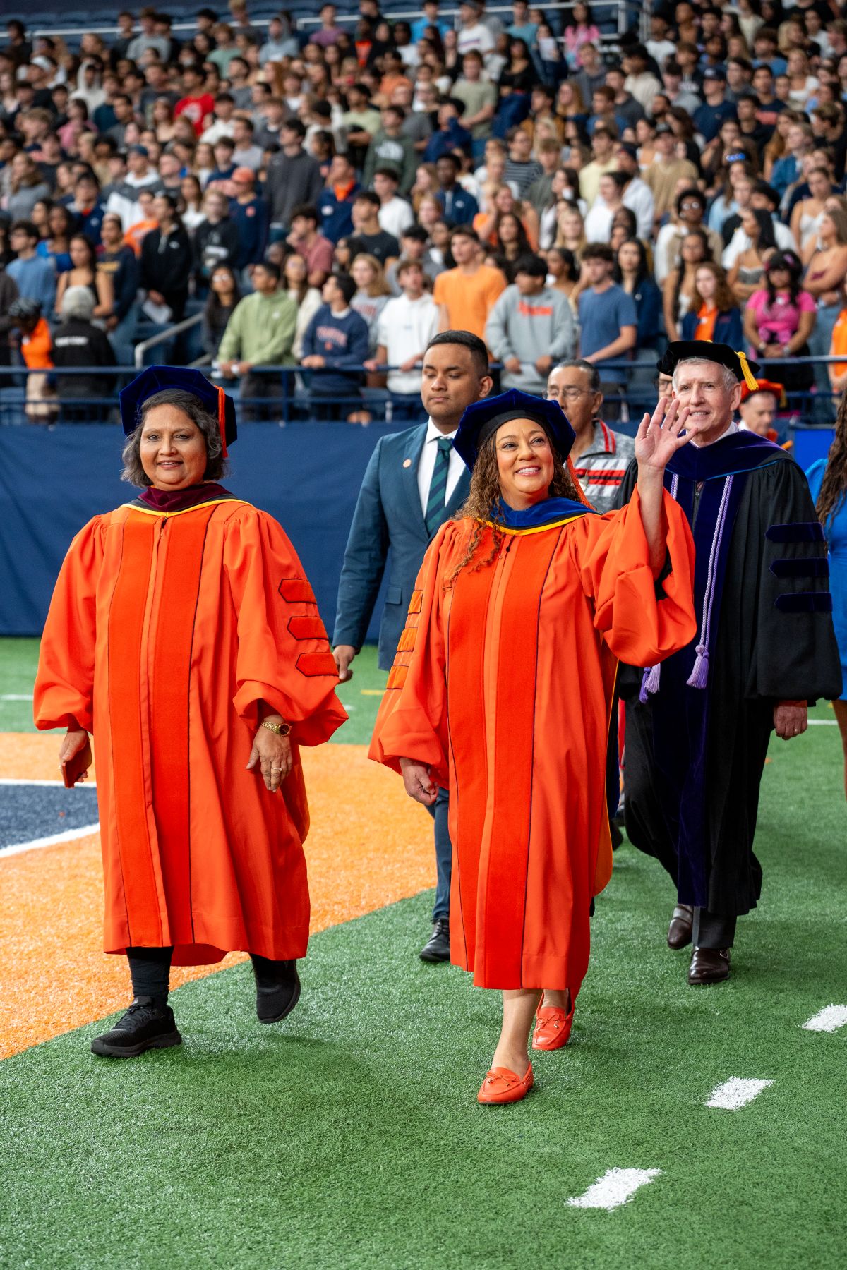 Community members in orange academic regalia walking across a field during a ceremony, with spectators in bleachers behind them.