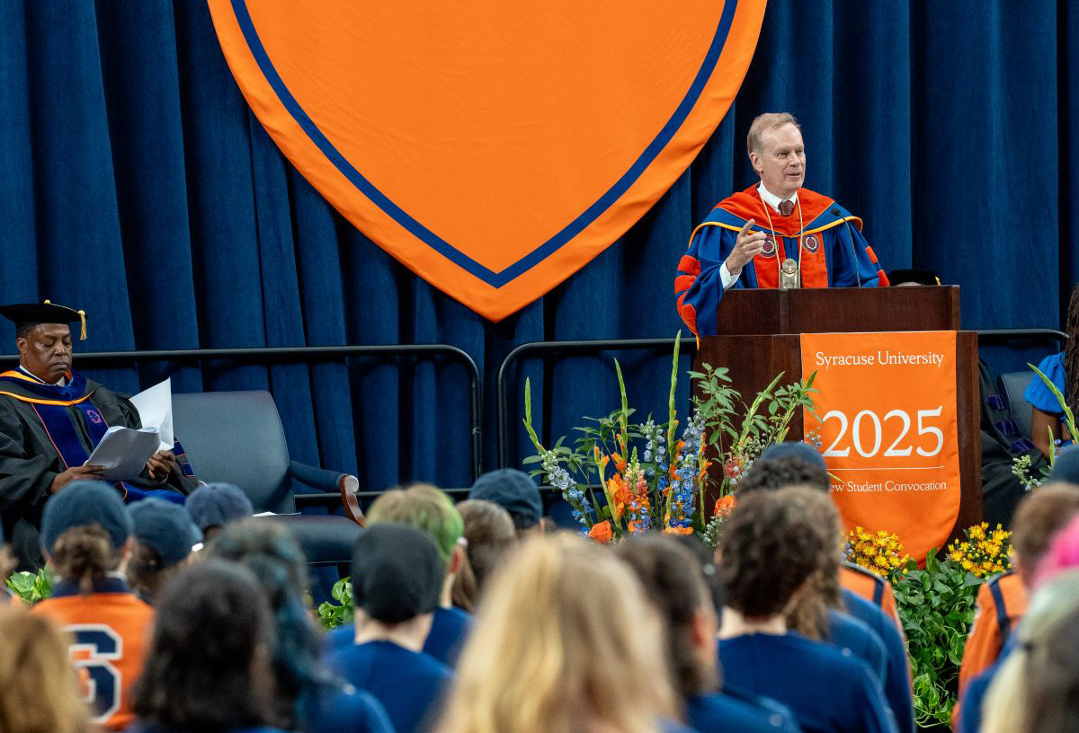 Chancellor Kent Syverud dressed in academic regalia at Syracuse University 2025 Student Convocation podium, with audience and orange banner in background.