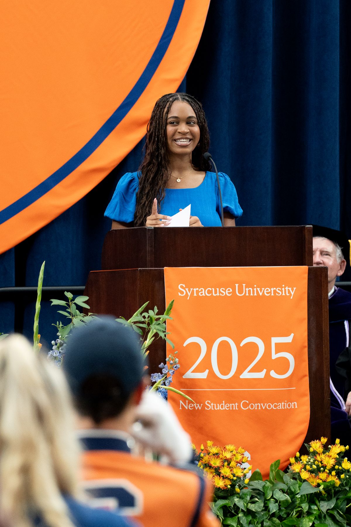 Speaker at podium with 'Syracuse University 2025 New Student Convocation' banner, in front of orange and blue backdrop and seated audience.