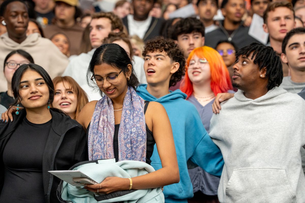 Group of people standing closely together, singing, in the JMA Wireless Dome at New Student Convocation. One person is seen holding a program.