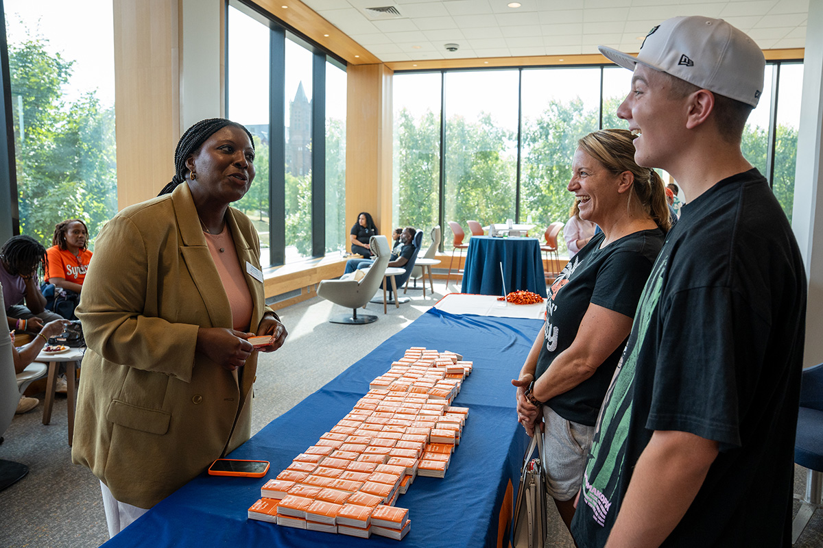 Person in tan blazer at a table with orange and white packages of tissues, engaging with two others in a sunlit room with seated attendees.