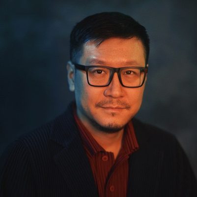Portrait of a man with black-rimmed glasses and facial hair wearing a burgundy shirt and dark sweater, photographed with warm lighting against a dark background.