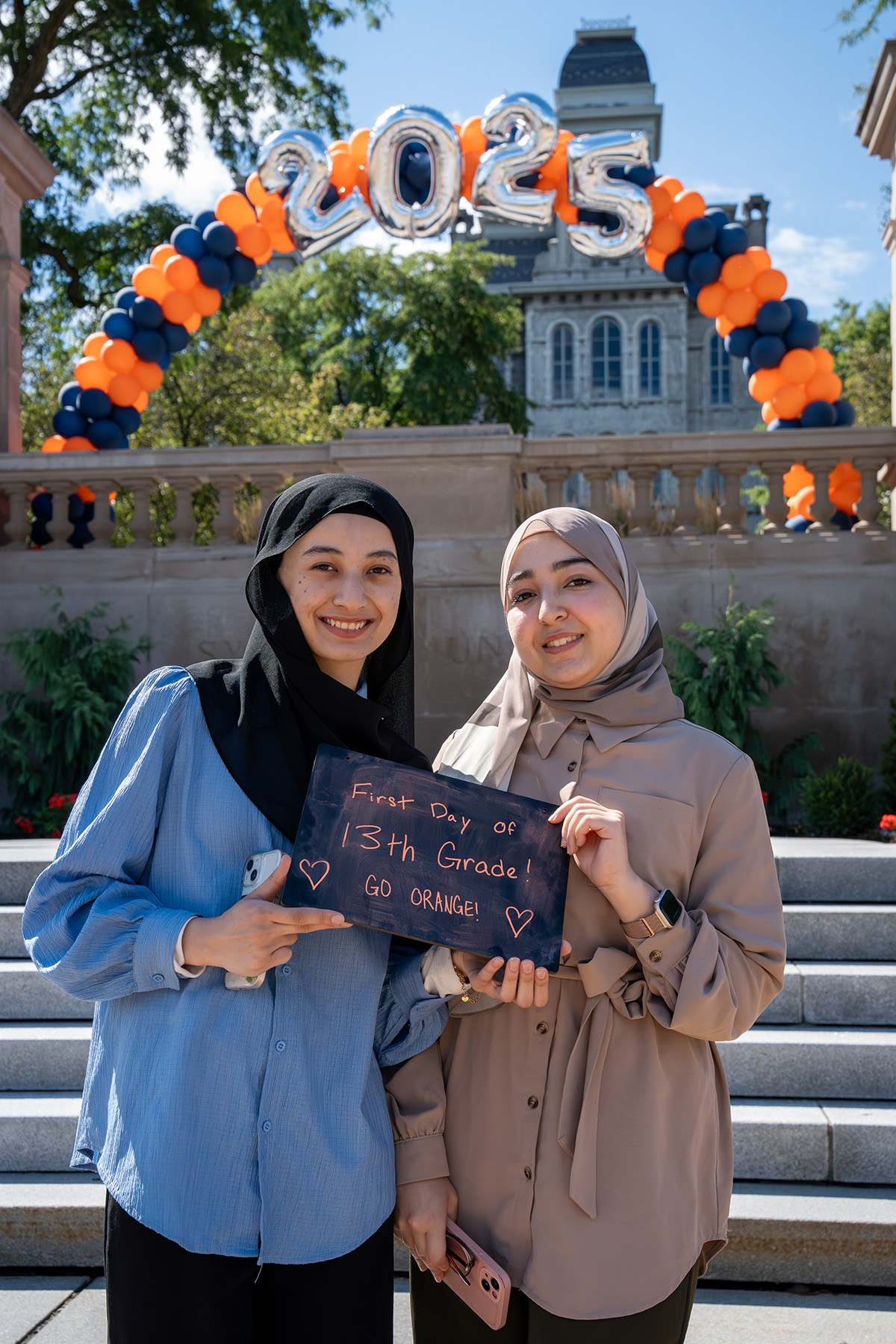 Two people holding a 'First Day of 13th Grade! Go Orange!' chalkboard sign under a balloon arch with '2025,' in front of the Hall of Languages.