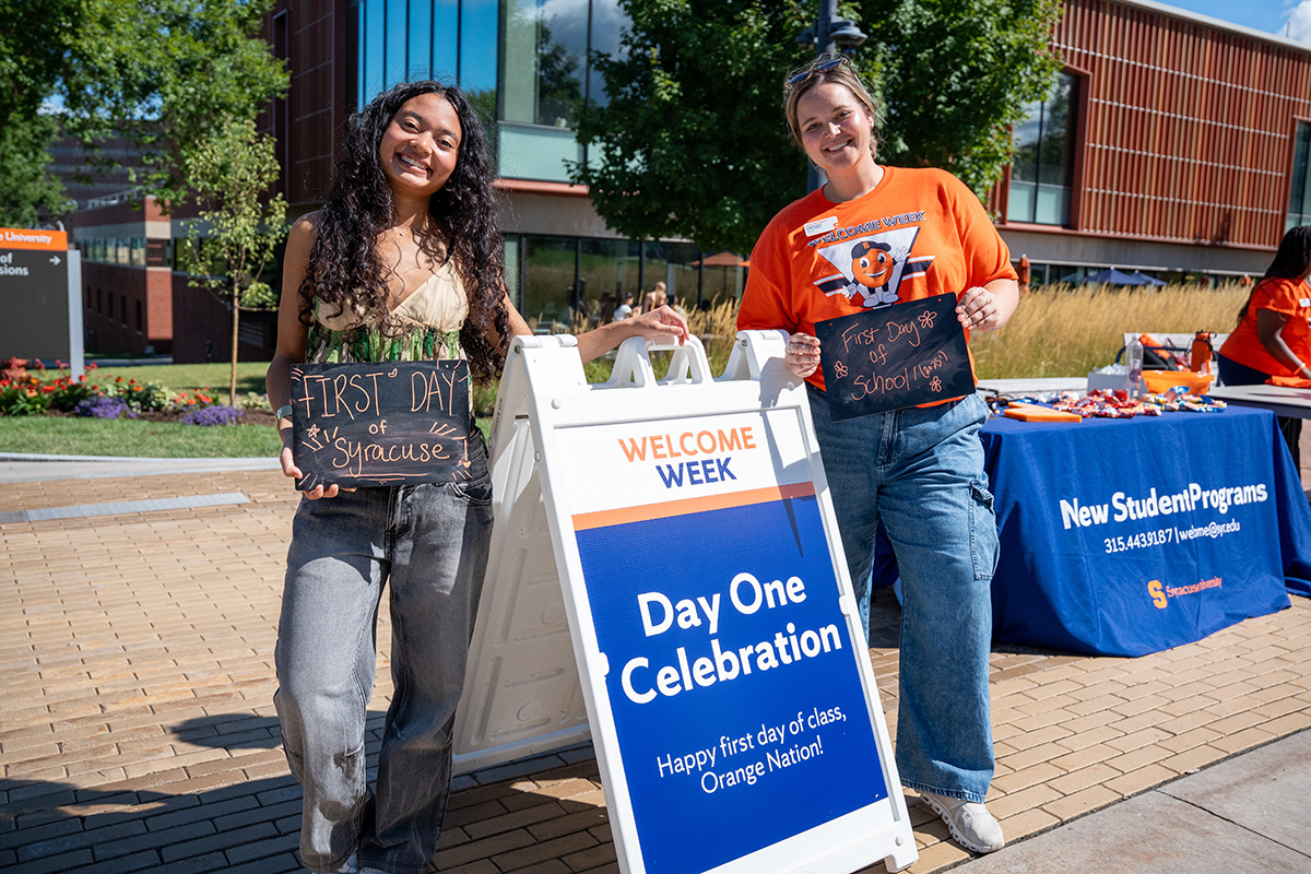 Two people holding 'First Day of Syracuse' and 'First Day of School' chalkboards beside a Welcome Week Day One Celebration sign, with a New Student Programs table in the background.