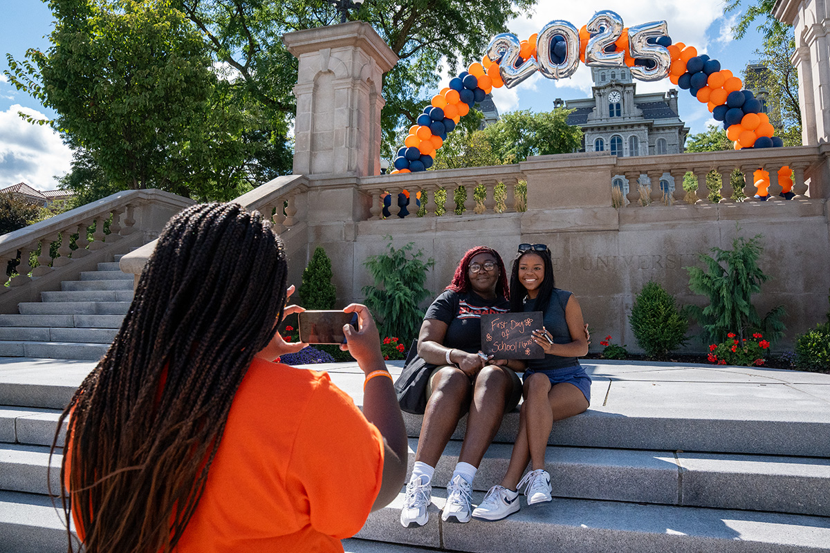 Two people holding a 'First Day of School' chalkboard sign under a balloon arch with '2025,' as someone in an orange shirt takes their photo on stone steps.