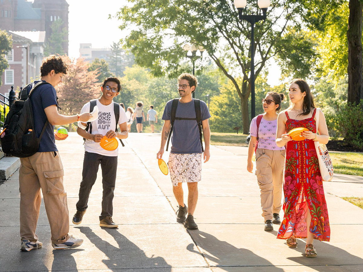 Five people walking on a sunny pathway with colorful frisbees, surrounded by trees and campus buildings.