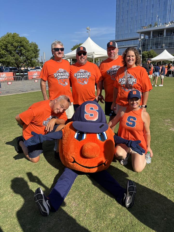A group of six Syracuse University fans in orange "Syracuse Orange" t-shirts pose with Otto the Orange, the university's mascot, at an outdoor campus event. Four adults stand in the back row while two people kneel beside the seated mascot, who wears his characteristic orange head, blue Syracuse cap, and blue sneakers on a sunny day with white tents and a modern building visible in the background.