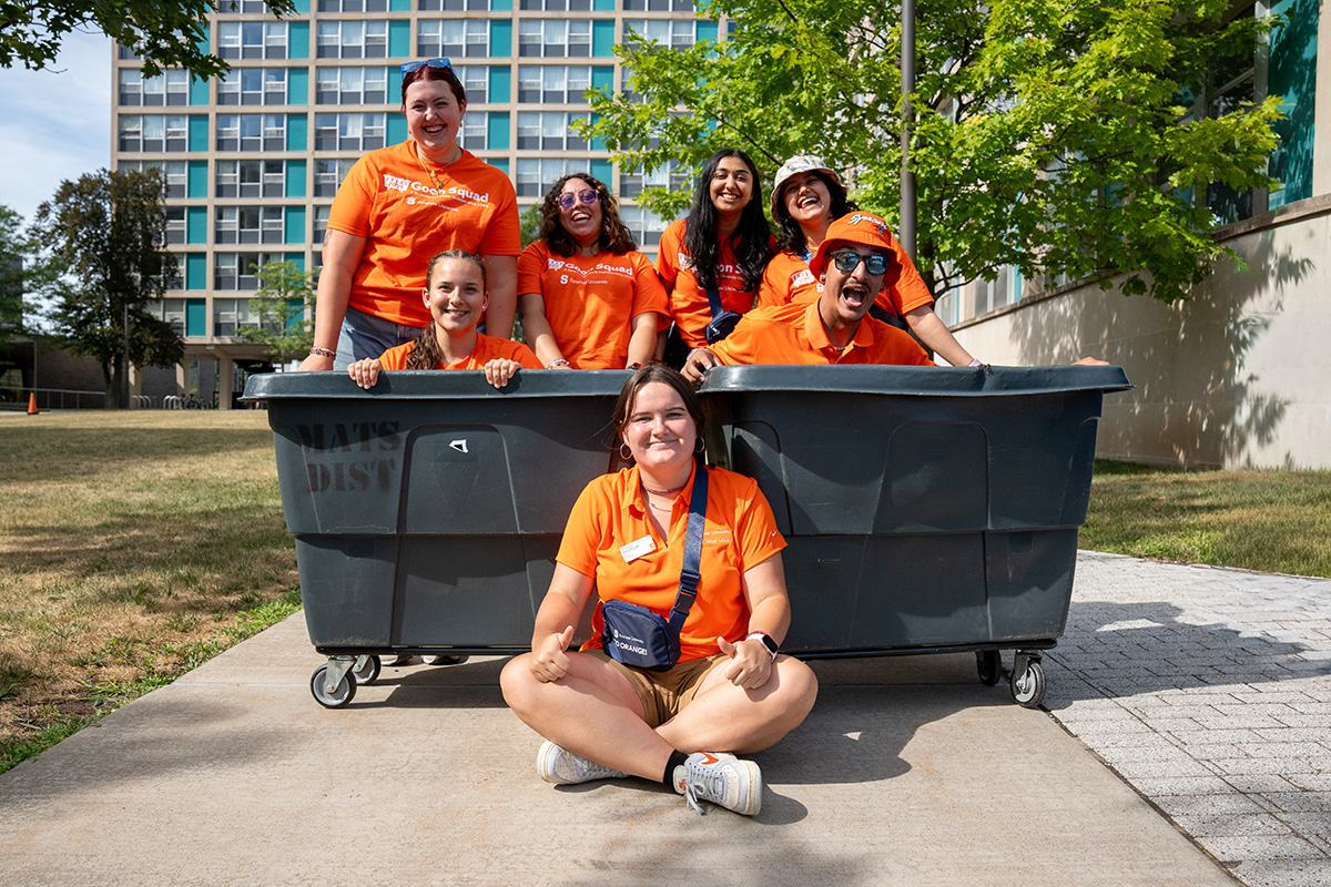 Seven people in matching orange shirts pose outdoors around a large black bin, with six people inside of it and one person sitting on the ground in front. A building, sidewalk and trees can be seen in the background.
