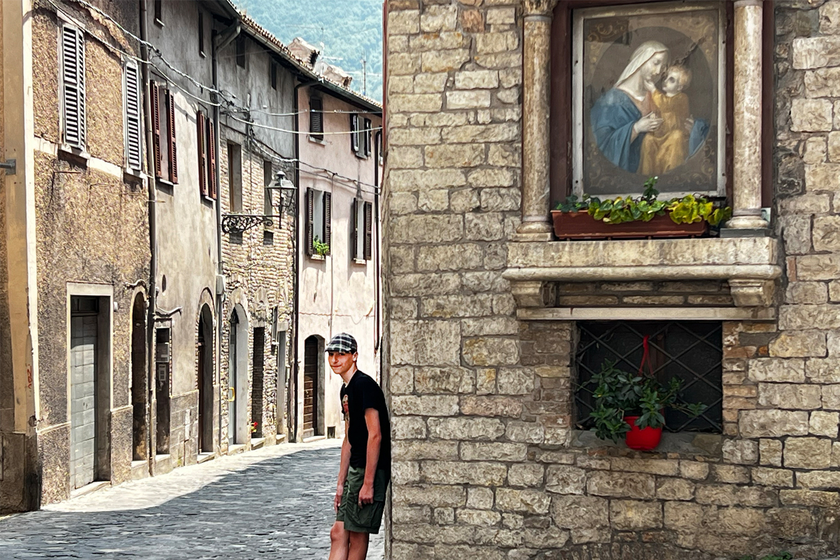 Man in cap and dark shirt walking down narrow cobblestone street in historic Mediterranean town, stone buildings with shuttered windows and religious artwork visible on wall.
