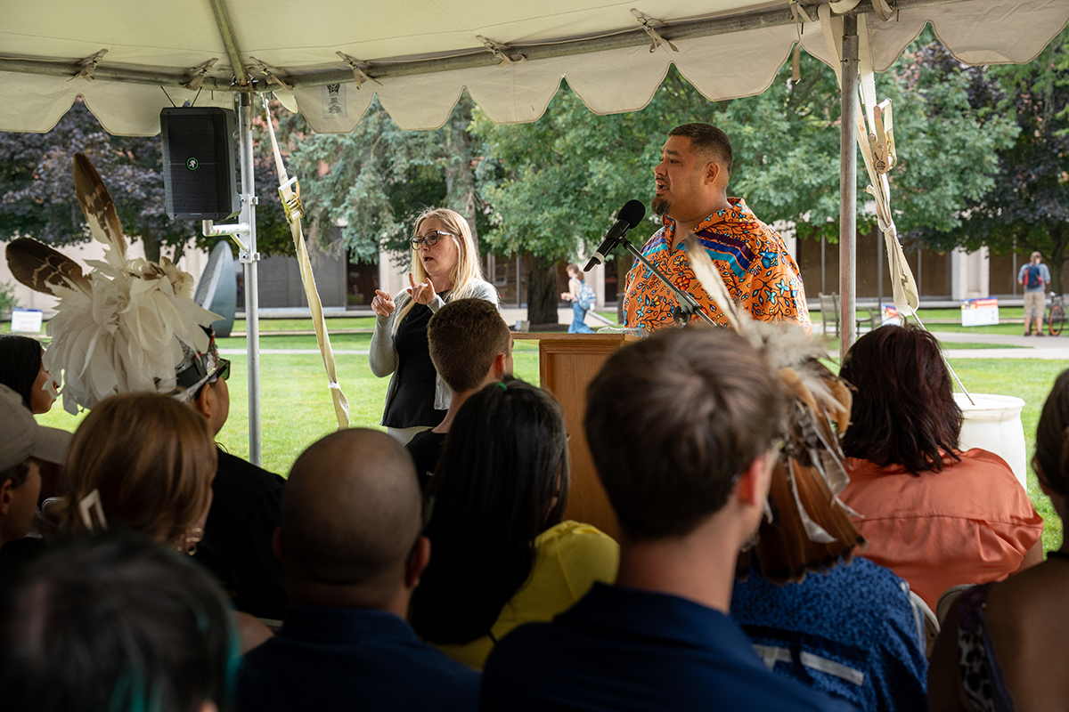 Audience seated under a tent listening to a speaker in a colorful shirt, accompanied by an ASL interpreter; trees and buildings in the background.