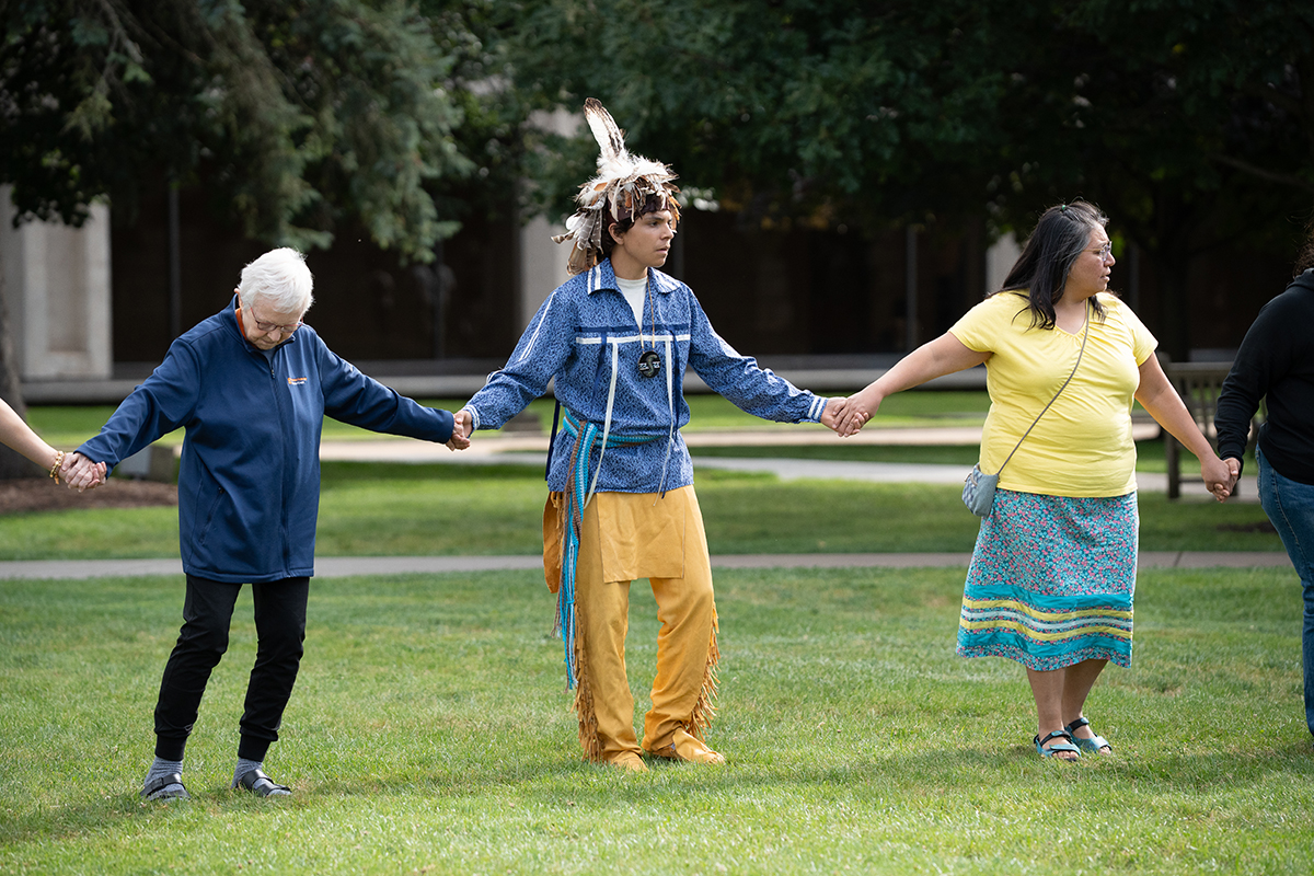 Group of people holding hands in a circle outdoors, including one person in traditional indigenous attire with a feathered headdress; grassy area with trees and buildings in the background.
