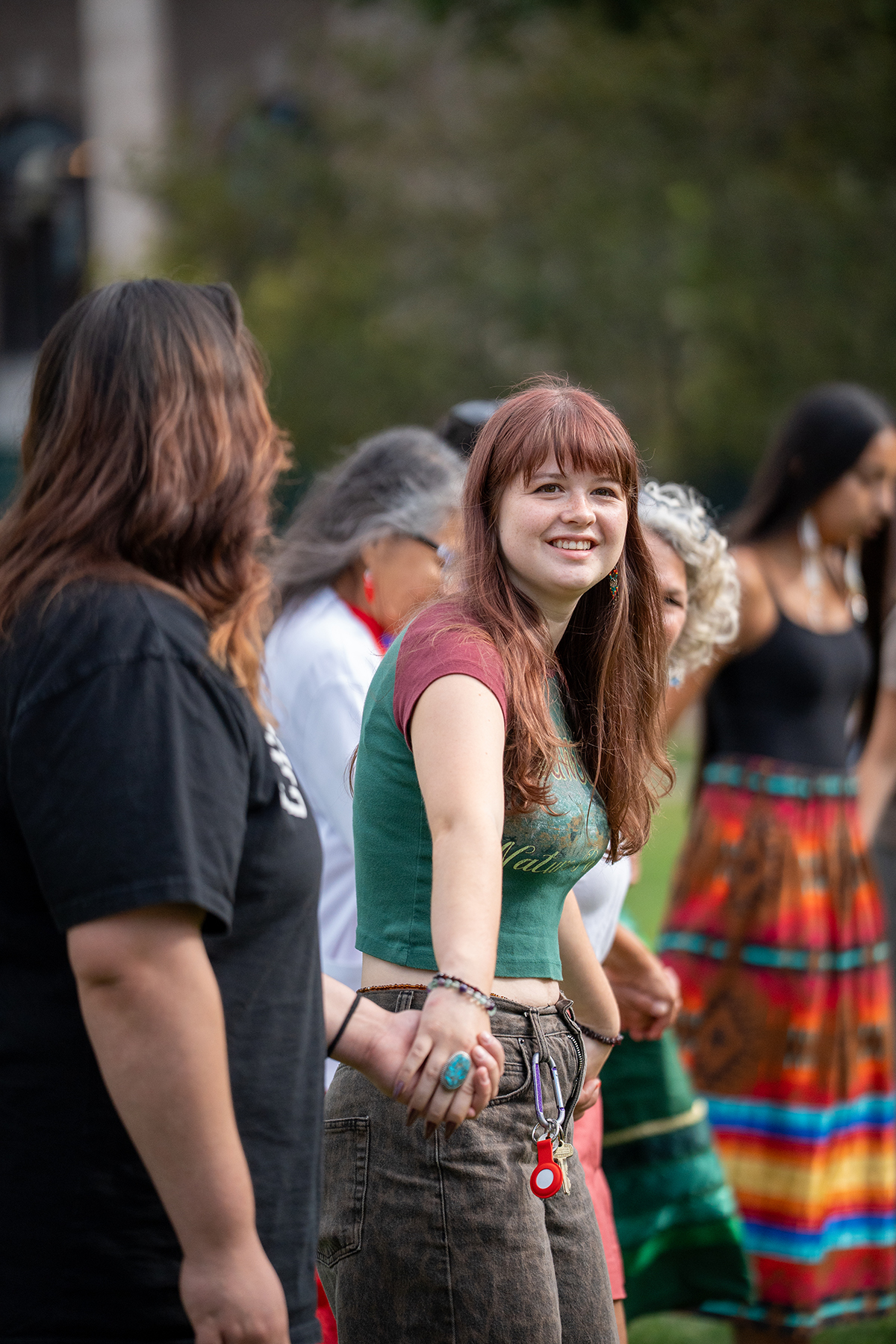 Group of people standing in a circle outdoors, holding hands in a grassy park with trees and buildings in the background.