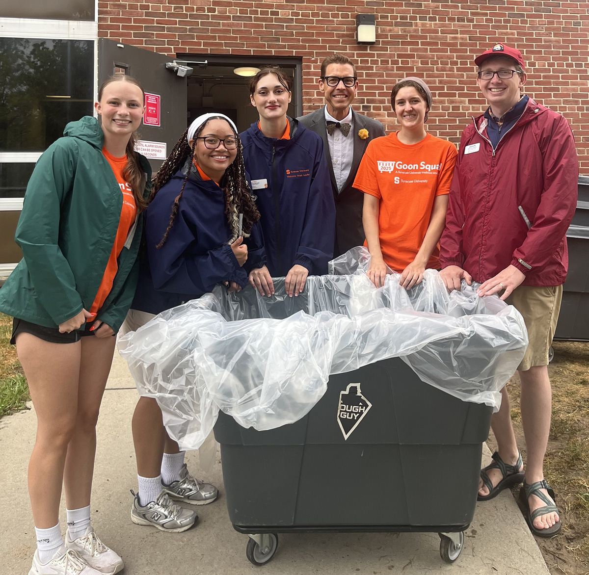 Six people gathered around a large black bin outside a brick building.