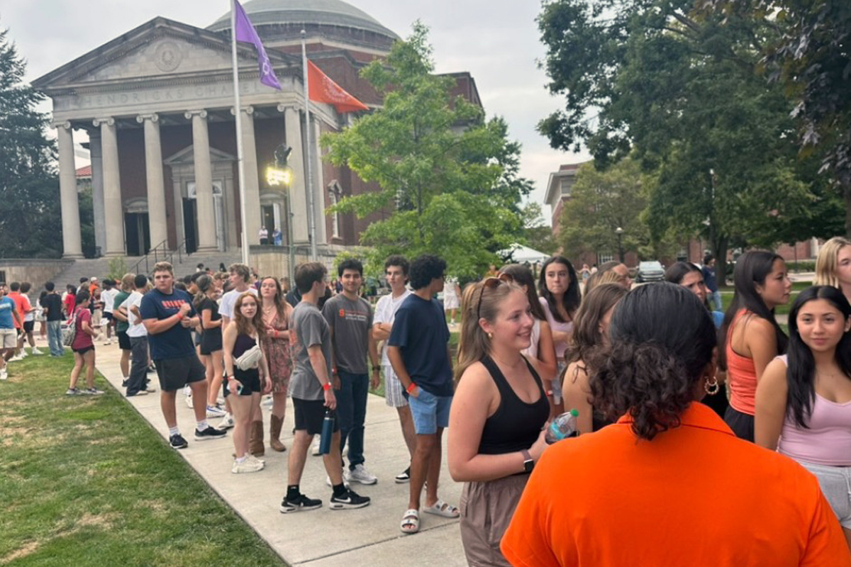 Large group of students gathered outdoors on a university campus near a neoclassical building with a dome, mingling and lining up during a casual event with school flags and university-themed attire.