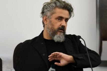Man with curly gray hair and beard speaking into a microphone, wearing a black shirt and gesturing with his hand.