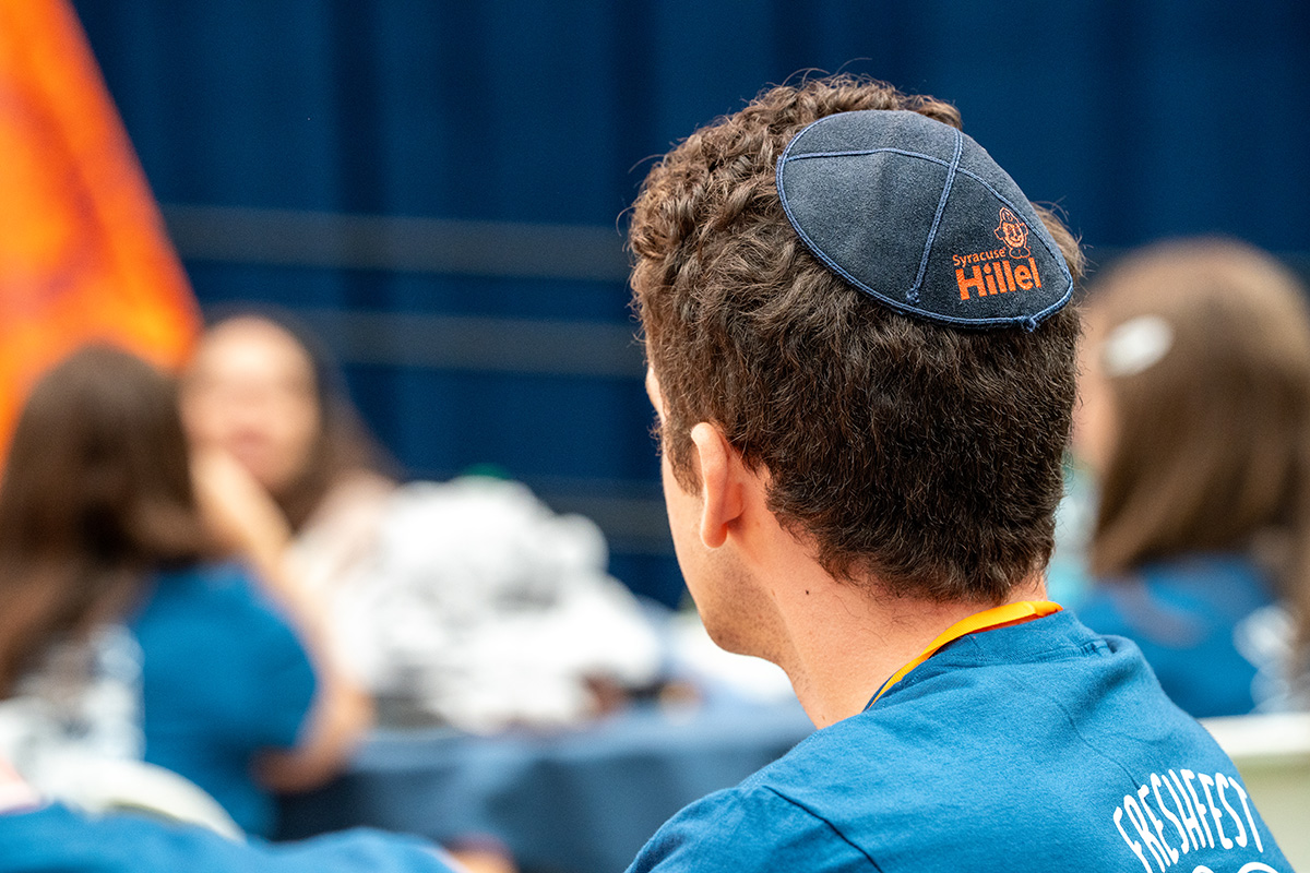 Person wearing a blue 'Syracuse Hillel' kippah and blue shirt, with others in similar attire blurred in the background.