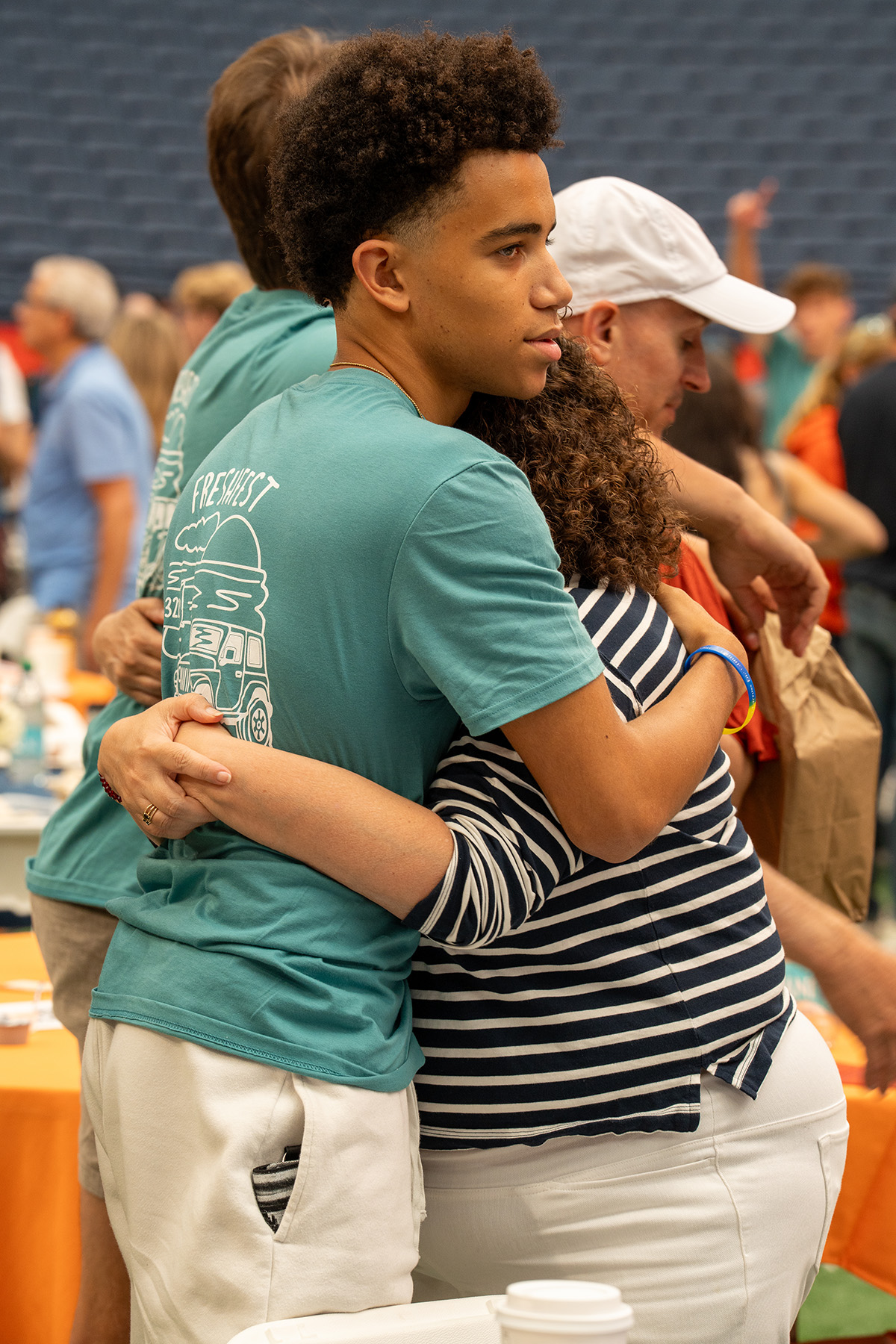 Two people embracing indoors, surrounded by others near tables with orange tablecloths and assorted items.