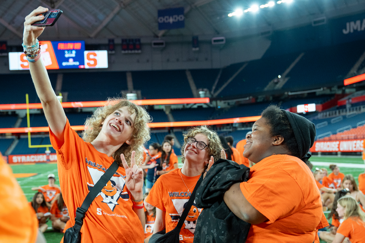 Three smiling people in orange Syracuse University shirts taking a selfie together inside a football stadium, with the JMA Wireless Dome's distinctive architecture and seating visible in the background.