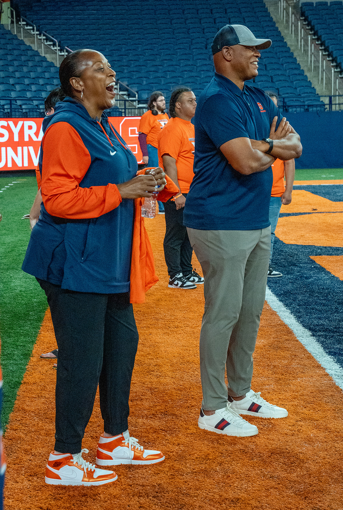 Two individuals standing on the sidelines of an indoor sports field, with Syracuse University signage and people in orange shirts in the background.