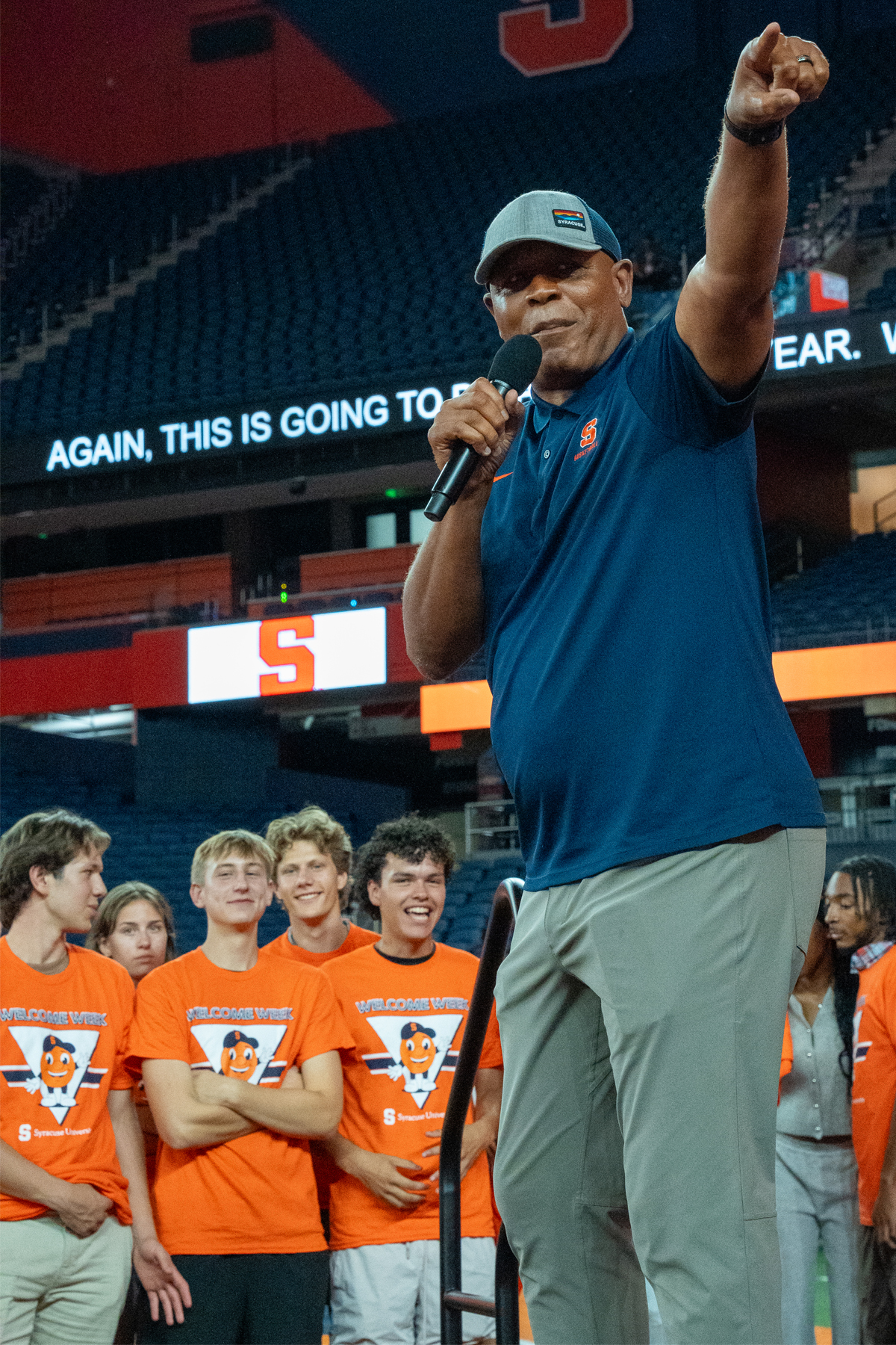 Man in blue Syracuse polo shirt and baseball cap speaking into microphone with raised fist while standing in front of excited college students wearing orange "Welcome Week" t-shirts in a stadium setting with Syracuse "S" logo visible in background.