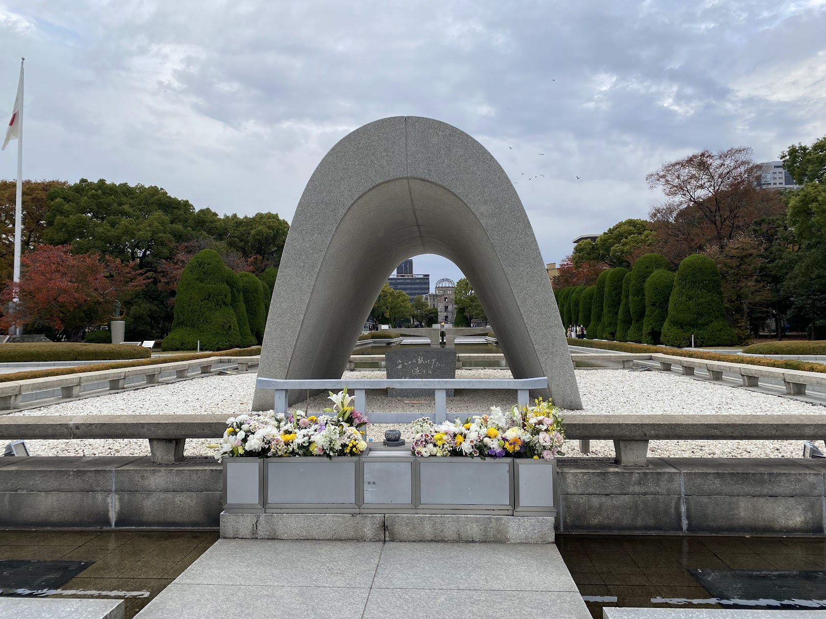 The Hiroshima Victims Memorial Cenotaph in Peace Memorial Park, with fresh flower offerings in front and the Atomic Bomb Dome framed through its arch in the distance.