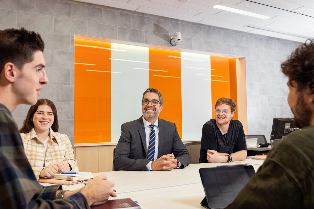 Five people, including a suited man and casually dressed individuals, sit around a table in a bright, modern room with orange and white wall panels, engaged in a collaborative discussion with notebooks and a tablet.