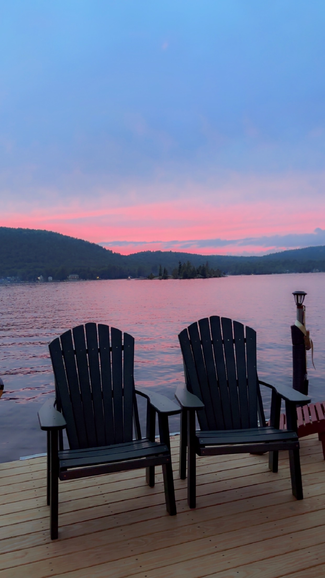 Two black Adirondack chairs on a wooden dock overlooking a calm lake at sunset, with pink and blue sky colors reflecting on the water and forested hills in the background.