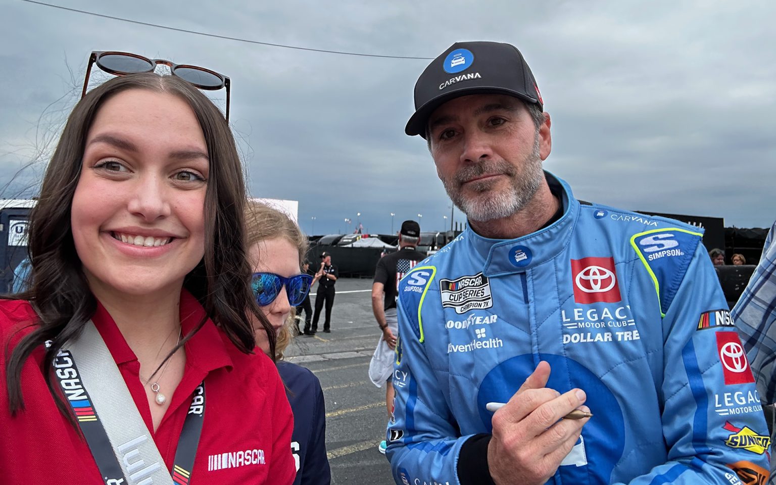 Young woman in red NASCAR polo shirt and lanyard posing for selfie with NASCAR driver in blue racing suit and Carvana cap at racing venue with overcast sky and race cars in background.