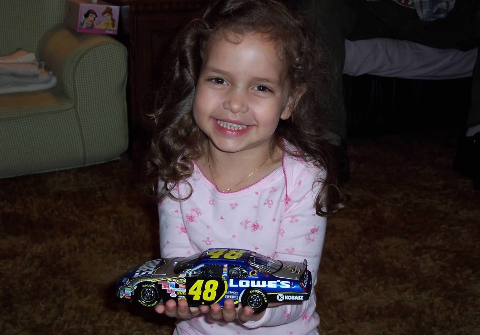 Young girl with curly brown hair wearing a pink floral pajama top, smiling while holding a blue and yellow #48 NASCAR die-cast race car toy in a home setting.