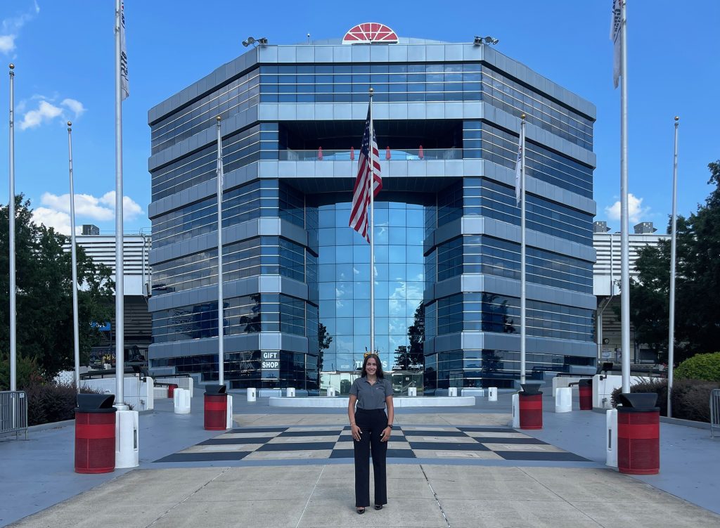 Woman in professional attire standing on a checkered plaza in front of a modern glass office building with an American flag, surrounded by flagpoles and landscaping under a blue sky with white clouds.