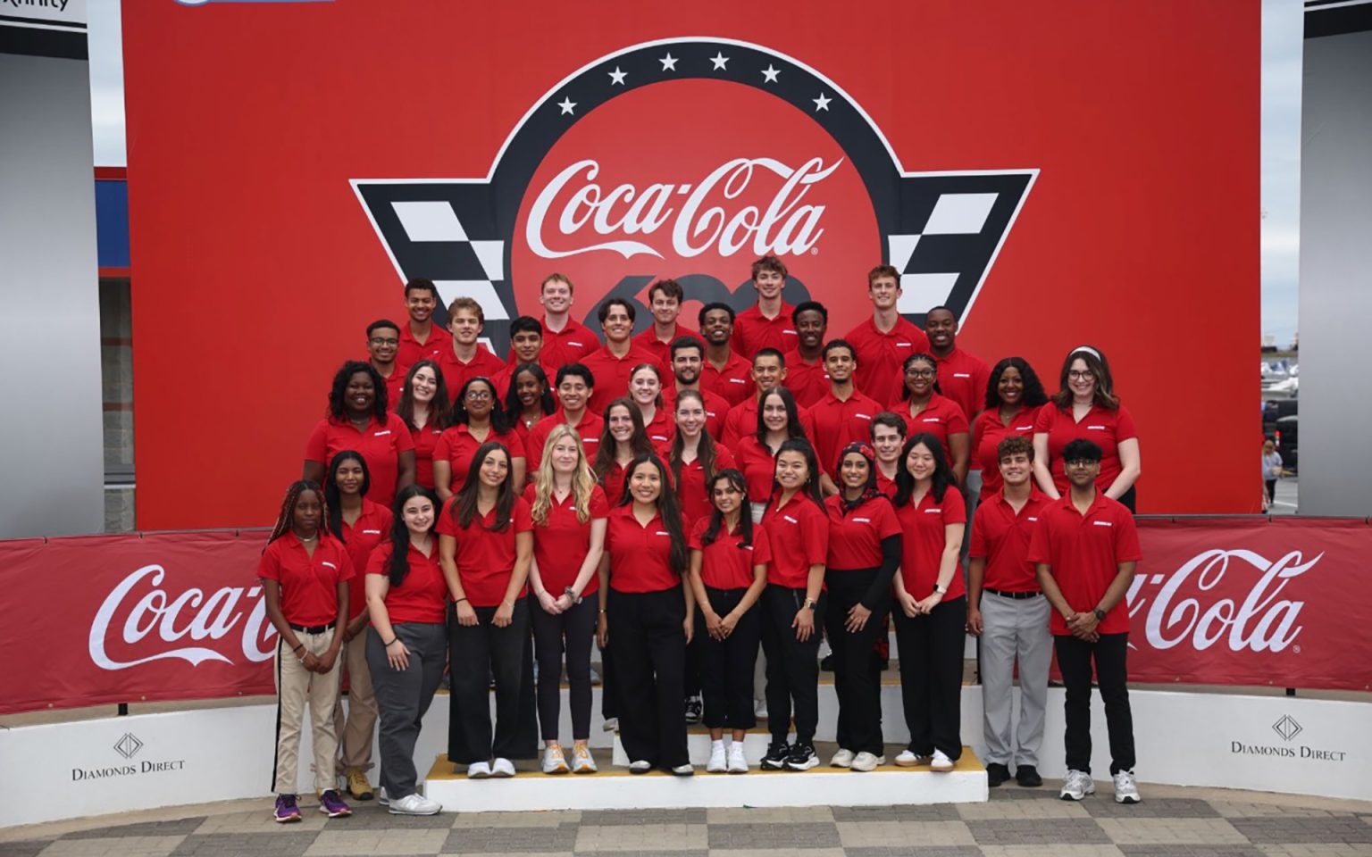 Large group of young people in matching red polo shirts posed together for a team photo in front of a prominent Coca-Cola branded backdrop with checkered flag design at what appears to be a racing venue or corporate event.