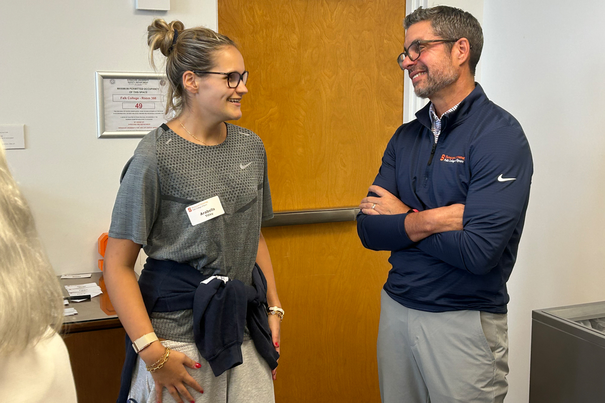 Two individuals, both wearing glasses, converse casually indoors near a wooden door; one wears a gray Nike t-shirt, the other a navy quarter-zip pullover and light gray pants.