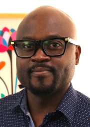A person wearing bold black glasses and a dark blue shirt with white polka dots standing in front of colorful abstract artwork