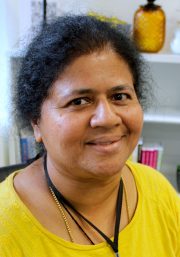 A person wearing a bright yellow top and a black lanyard seated in front of a shelf containing books and decorative items, including a yellow vase