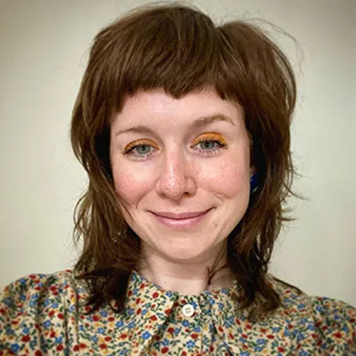 Portrait of a woman with brown layered hair and bangs, wearing a floral patterned collared shirt, smiling at the camera against a light neutral background.