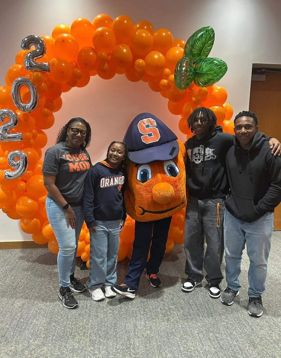 Four people pose indoors with Otto, Syracuse University's mascot, in front of a festive balloon arch featuring "2029" in silver balloons and leafy green accents, celebrating school spirit and a future graduating class.