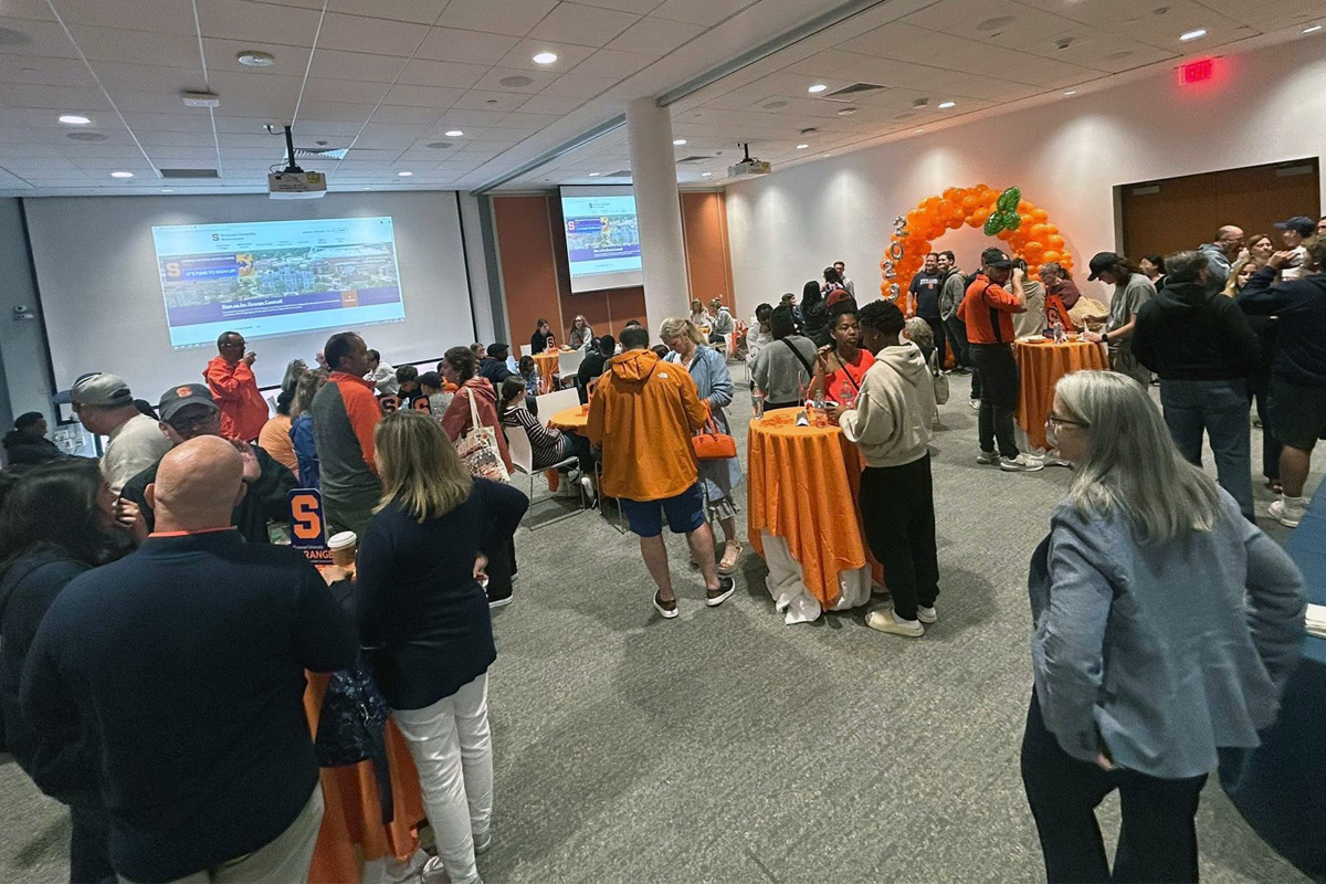 Attendees gather around orange-covered tables in a modern, well-lit room for a Syracuse University event, with festive balloon decorations and a presentation screen displaying university branding.