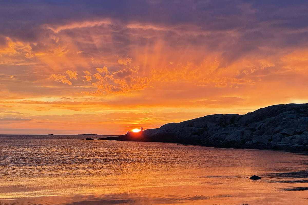 Golden sunset over calm water with dramatic orange and purple clouds reflected on the surface, silhouetted rocky coastline in background.