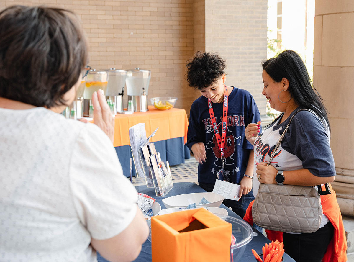 Three people at a table with informational materials and an orange box, with snacks and drinks in the background.