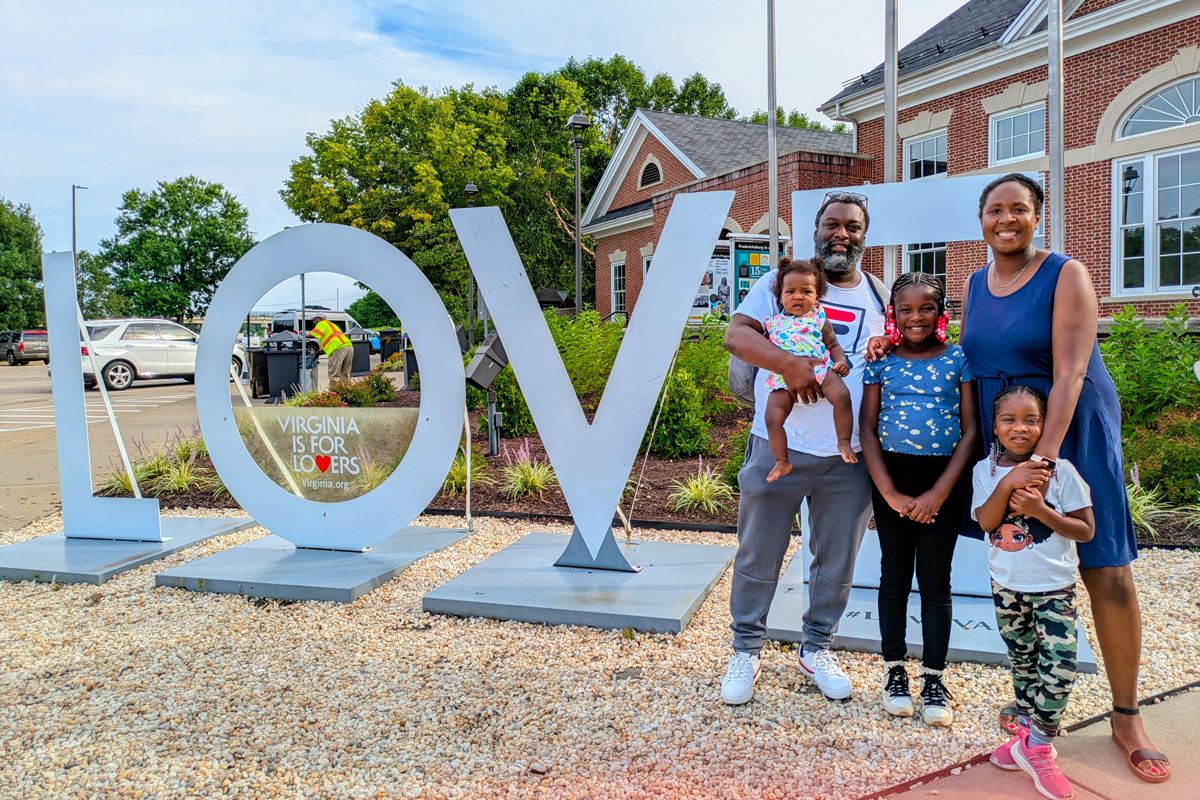 Family of five posing beside large white 'LOV' letters sculpture with 'Virginia is for Lovers' sign, brick buildings and trees in background on sunny day.