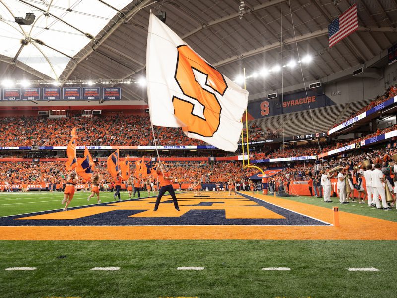 Cheerleaders wave flags on a football field as fans in orange fill the JMA Wireless Dome, with a large white flag bearing an orange 'S' in the foreground.