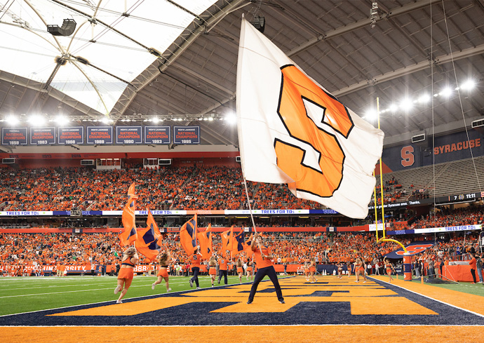Cheerleaders wave flags on a football field as fans in orange fill the JMA Wireless Dome, with a large white flag bearing an orange 'S' in the foreground.