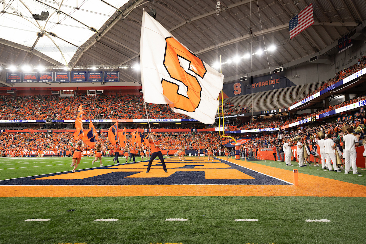 Cheerleaders wave flags on a football field as fans in orange fill the JMA Wireless Dome, with a large white flag bearing an orange 'S' in the foreground.